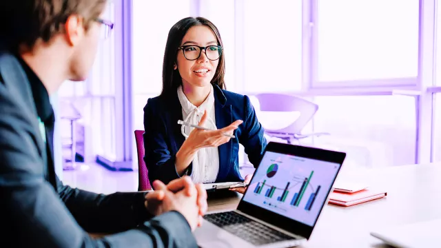 Young female manager talking to colleagues gesture and smiling having consultancy meeting about business, positive woman in formal wear asking question while man sitting near laptop with infographic