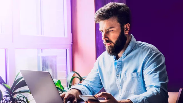 Image of man at the desk, looking at the laptop and holding a smartphone in the left hand