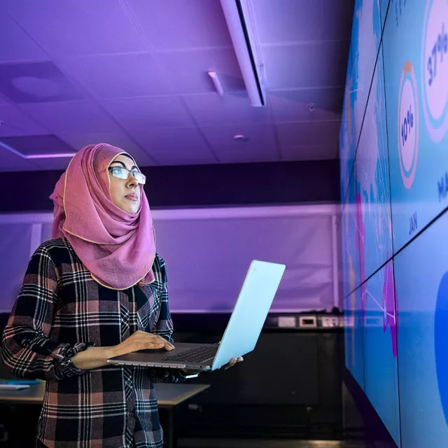 Image of woman holding a laptop and analyzing data displayed on large screens