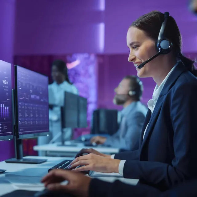 Professionals in a high-tech control room with purple lighting, working at computer monitors displaying data analytics and graphs, wearing headsets for communication and monitoring.