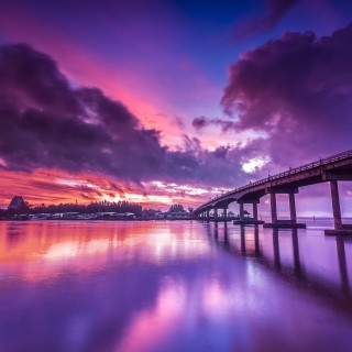 Serene sunset scene with a bridge over calm water, vibrant sky colors, and city silhouettes.