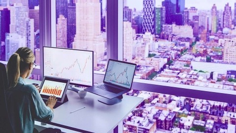 Image of a blonde woman at her office desk, analyzing data displayed on a tablet, a monitor and a laptop